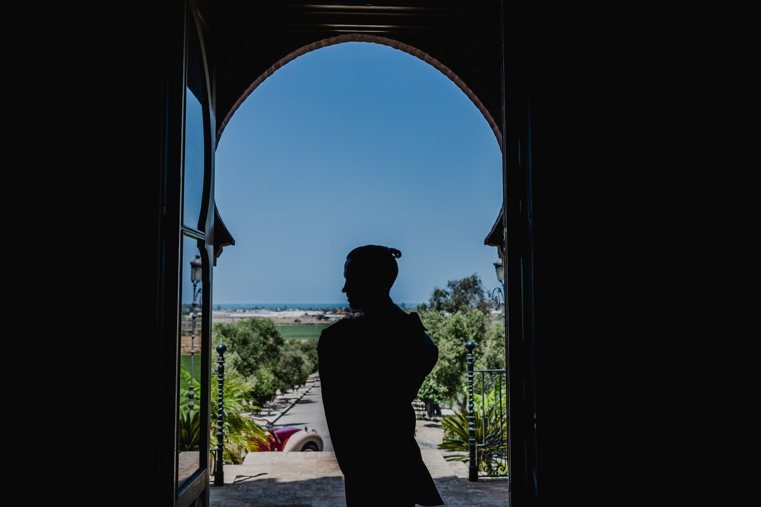 Silueta de una persona parada en una puerta arqueada, con vista a un paisaje de árboles verdes y cielo azul.