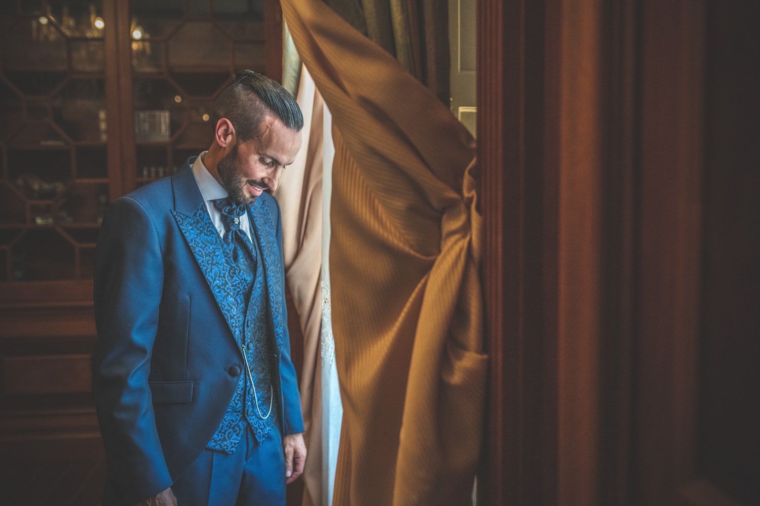Hombre con traje azul, mirando hacia abajo, de pie junto a una cortina en una habitación con una estantería.
