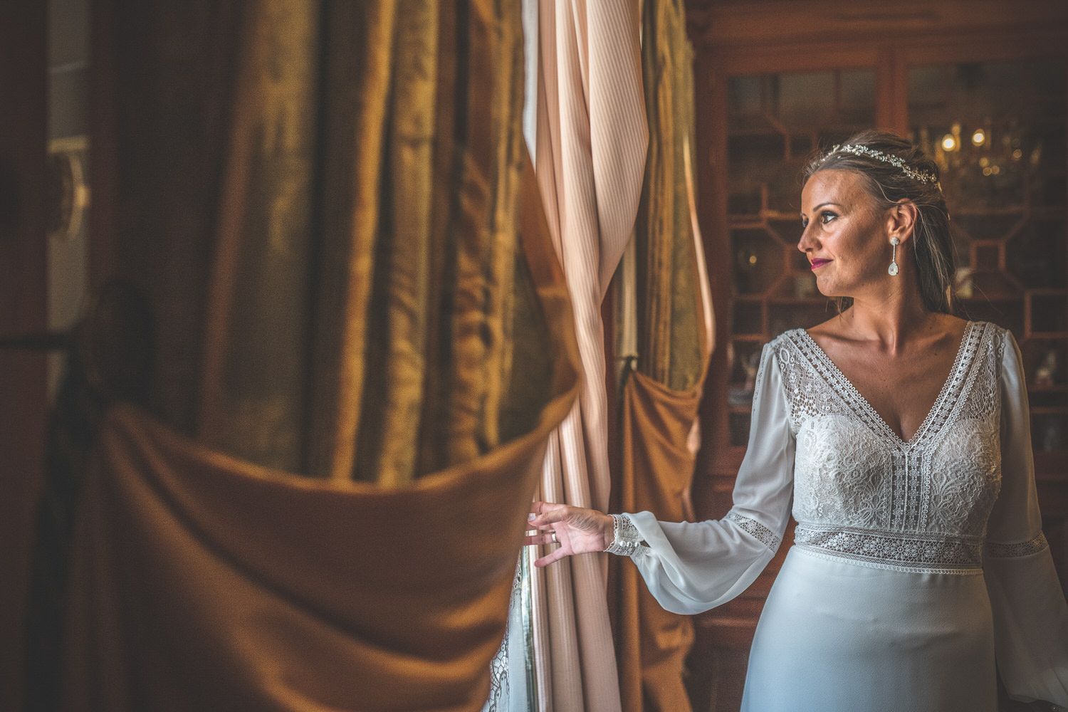 Una mujer con un vestido de novia blanco mira por una ventana, con una cortina dorada a su izquierda.