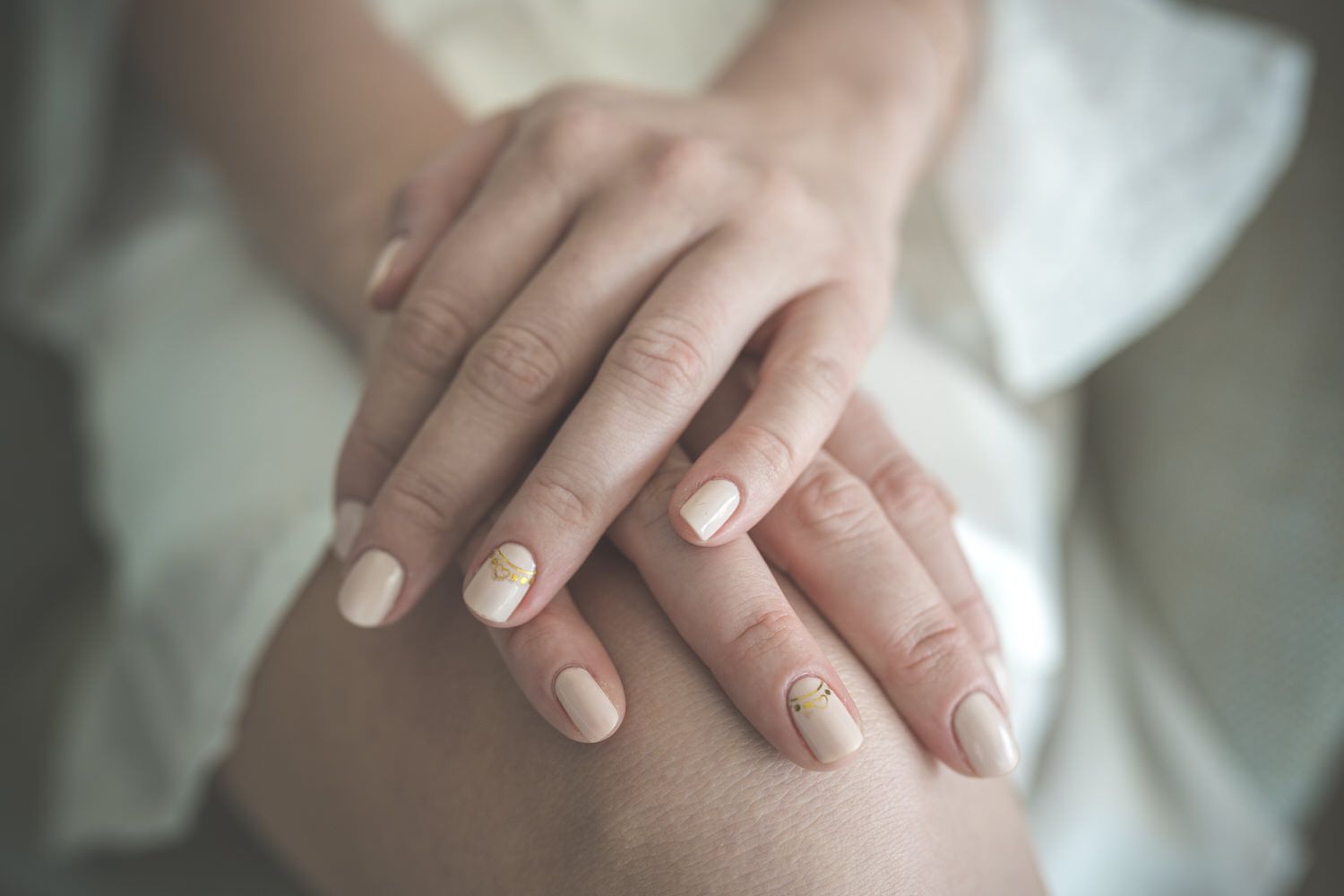 Manos juntas sobre el regazo, esmalte de uñas claro. Piel pálida, enfoque suave.