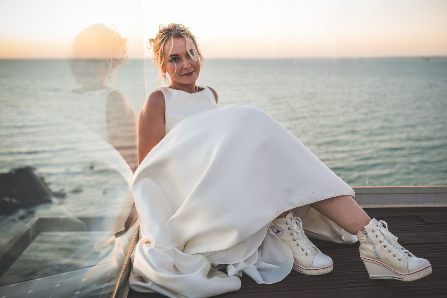 Una mujer con un vestido blanco y zapatillas de cuña se sienta en una terraza de madera con vistas al océano al atardecer.