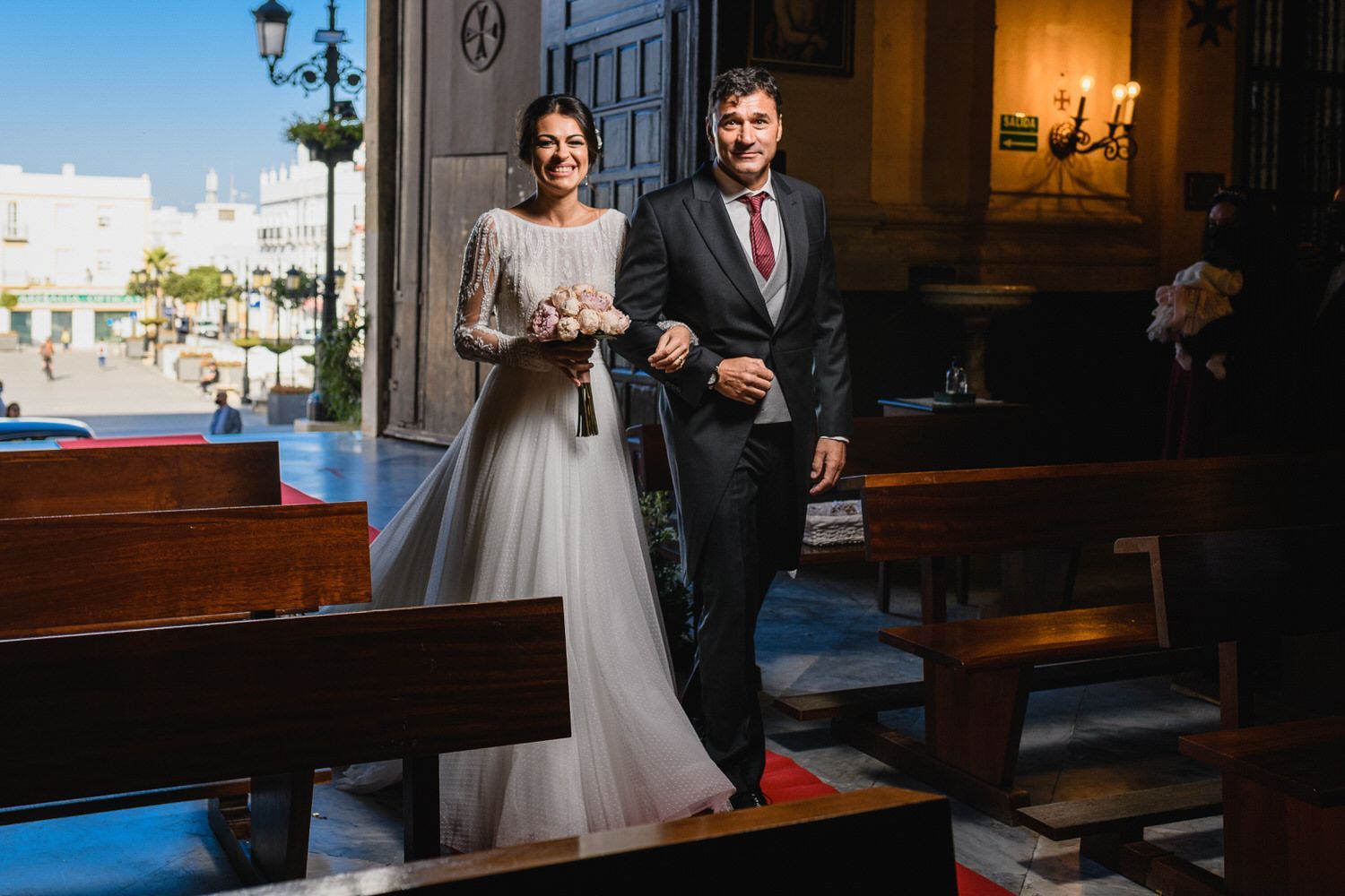 Los novios salen de una iglesia, sonriendo, cogidos del brazo. La novia con vestido blanco, el novio con traje.