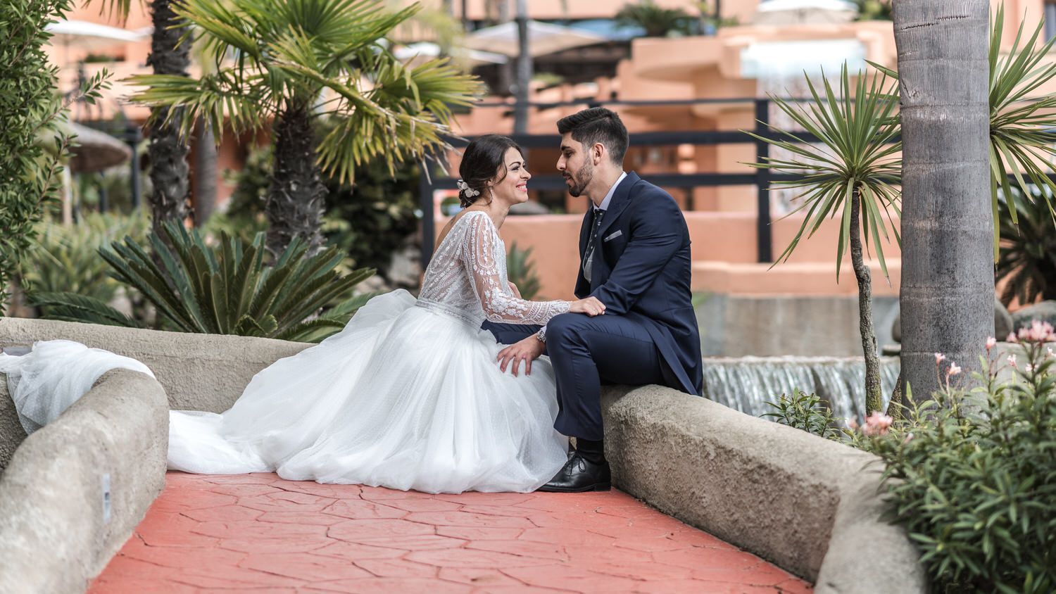 Los novios se miran sentados en un puente bajo. Ella lleva un vestido blanco, él un traje azul marino.