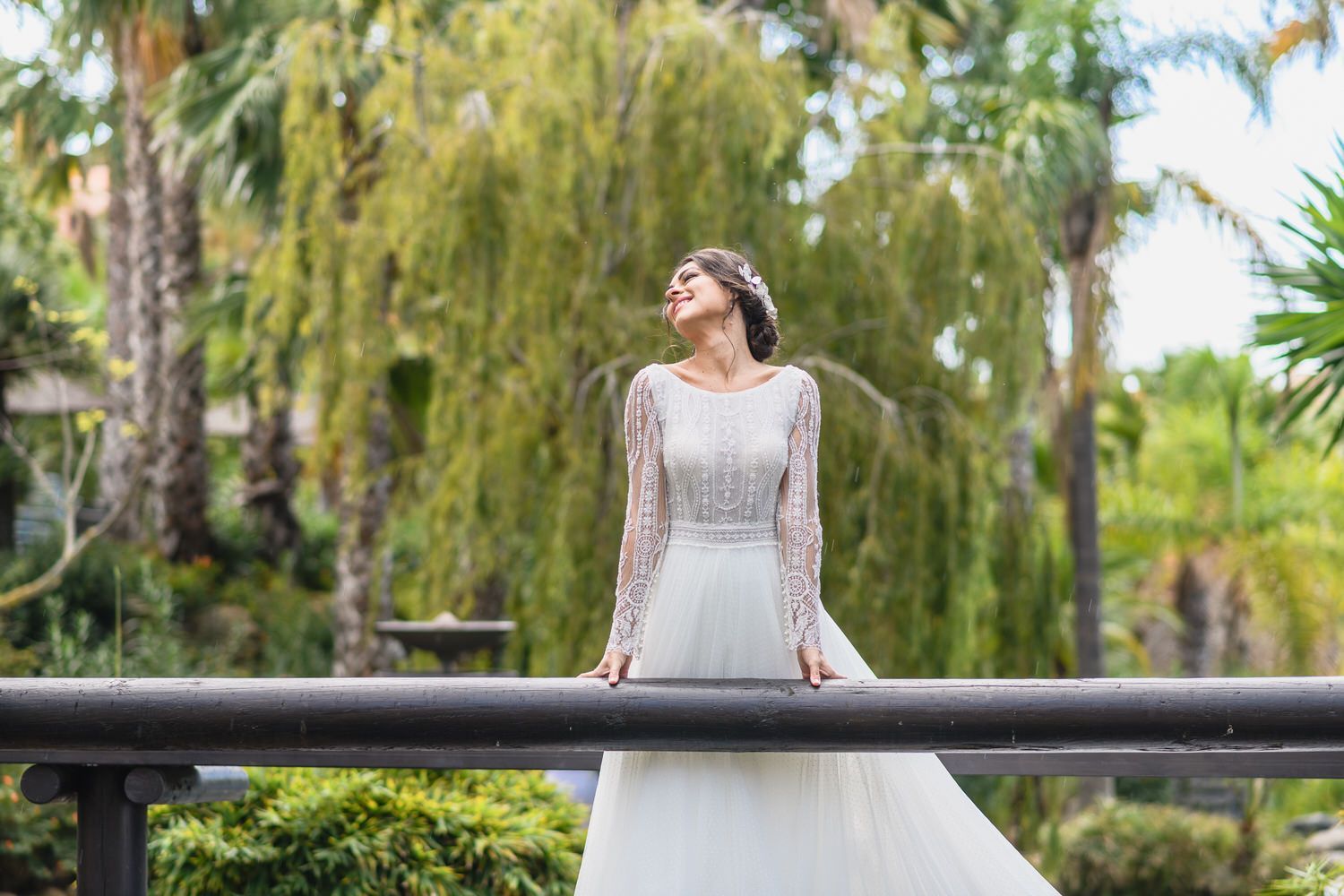 Novia con vestido blanco, brazos en la barandilla, sonriendo, ojos cerrados, entorno al aire libre con árboles.
