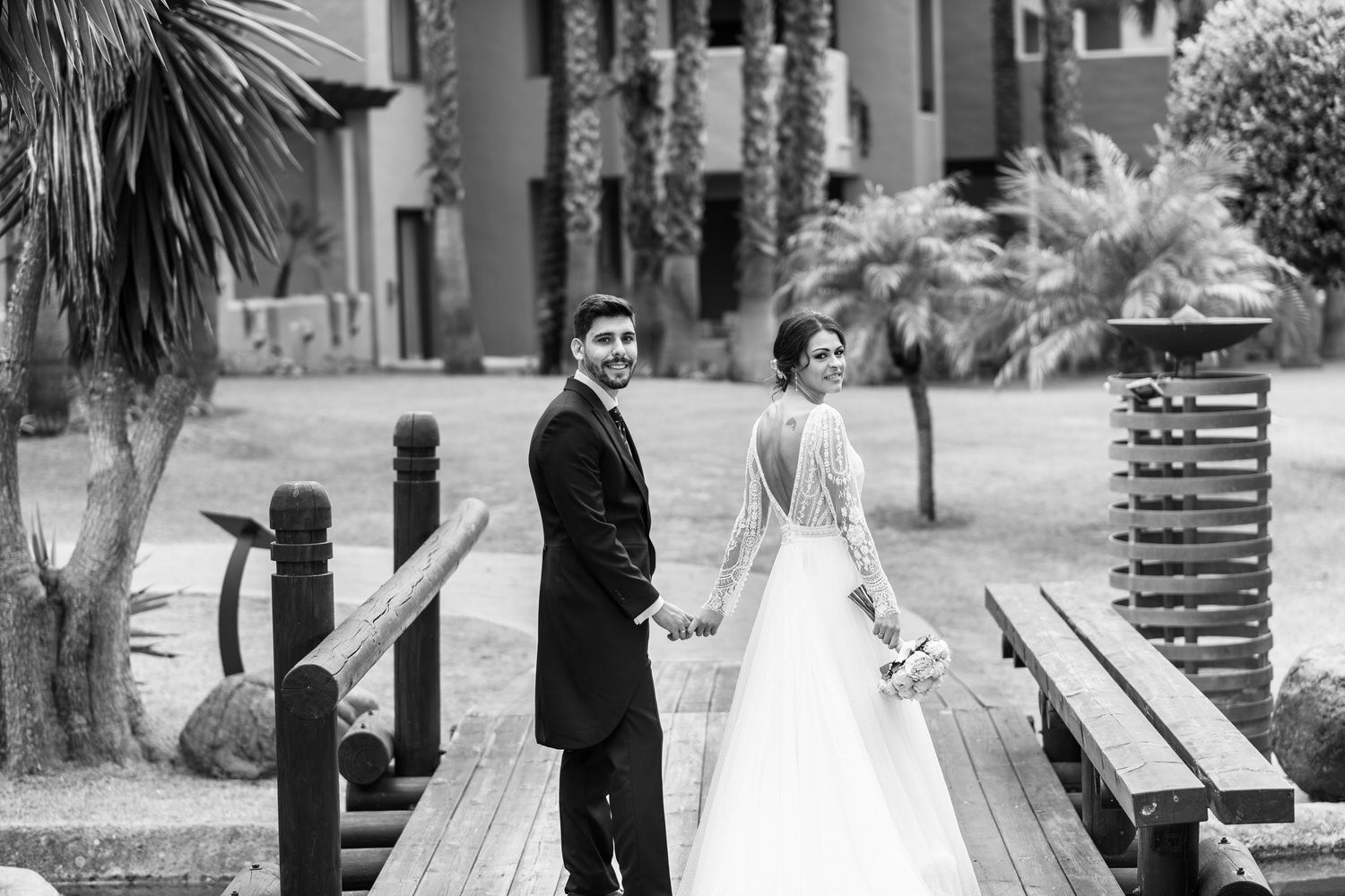 Recién casados ​​tomados de la mano, caminando sobre un puente de madera, mirando hacia la cámara.