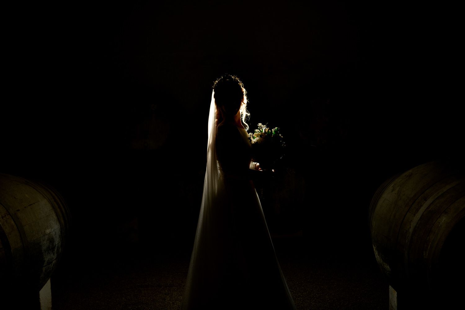 Silueta de una novia sosteniendo un ramo en una habitación poco iluminada con grandes barriles.