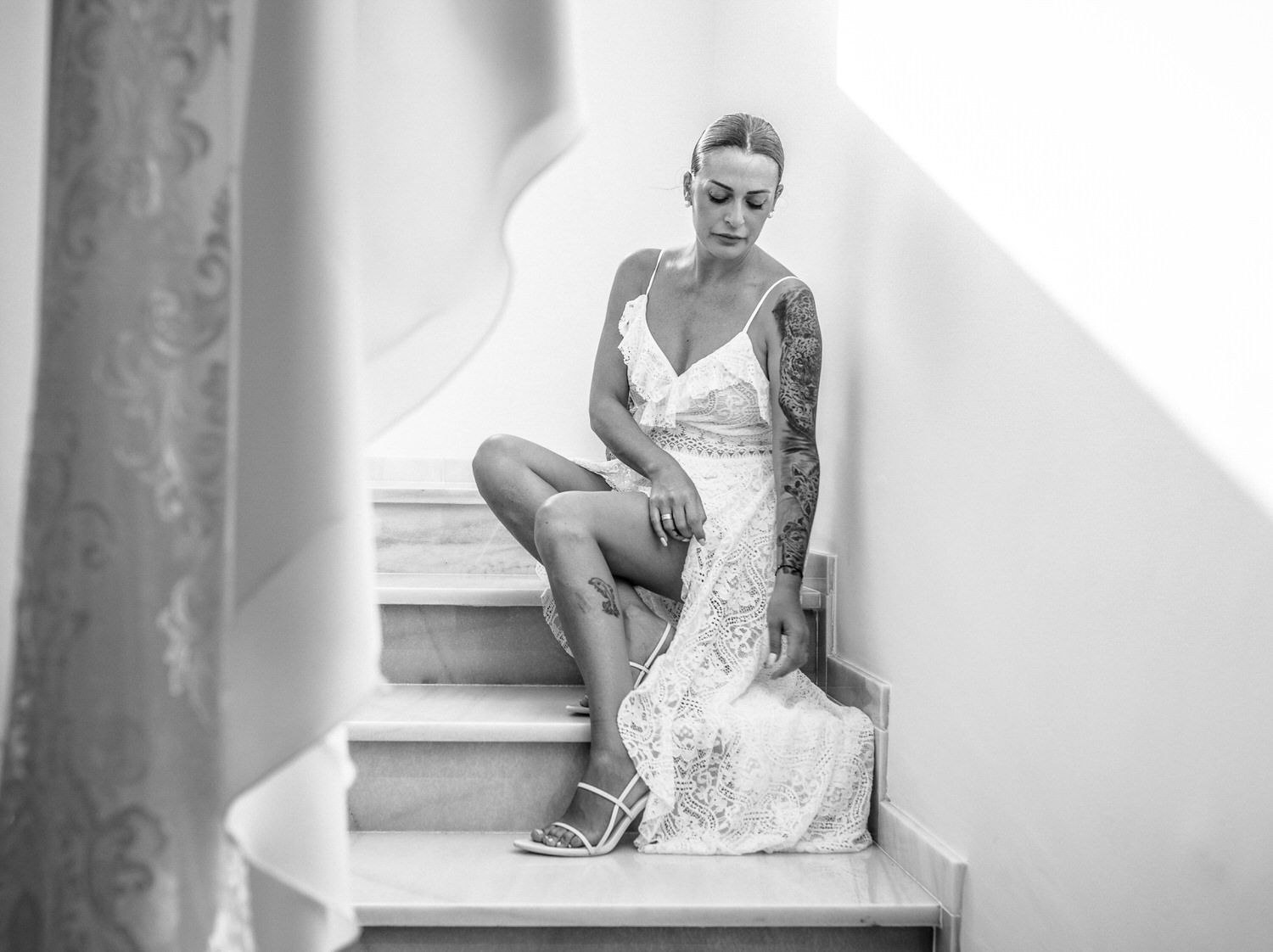 Mujer con vestido de encaje blanco sentada en las escaleras, mirando hacia abajo. Tatuaje visible en el brazo.