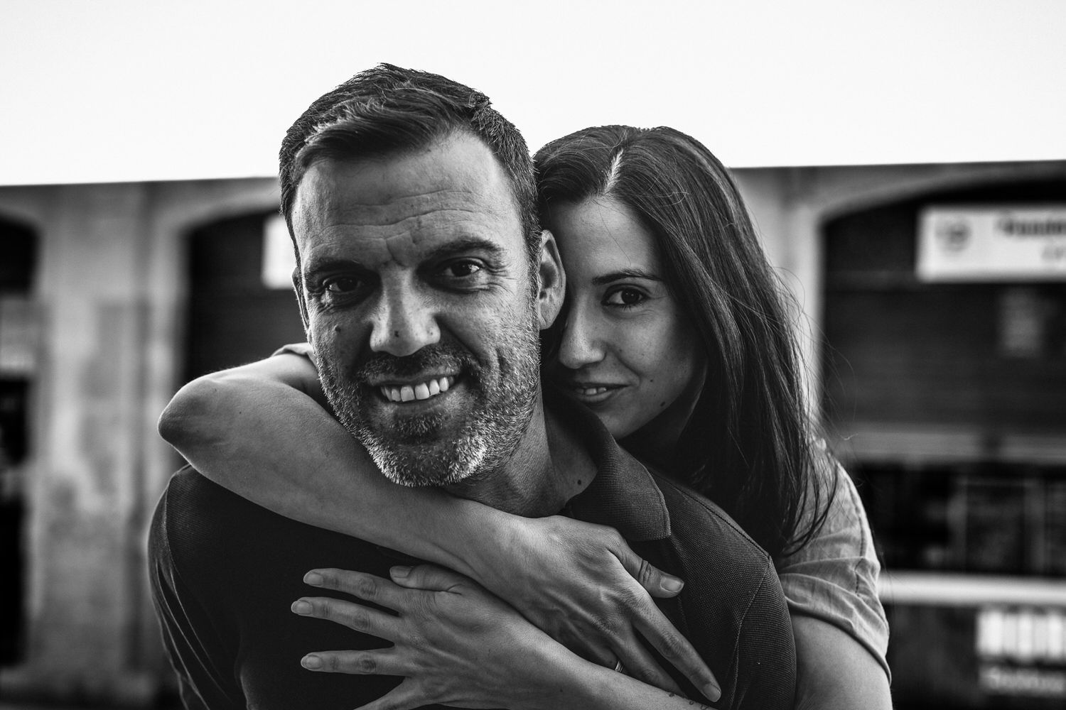 Una mujer abraza a un hombre sonriente; ambos miran a la cámara. Blanco y negro, exterior.