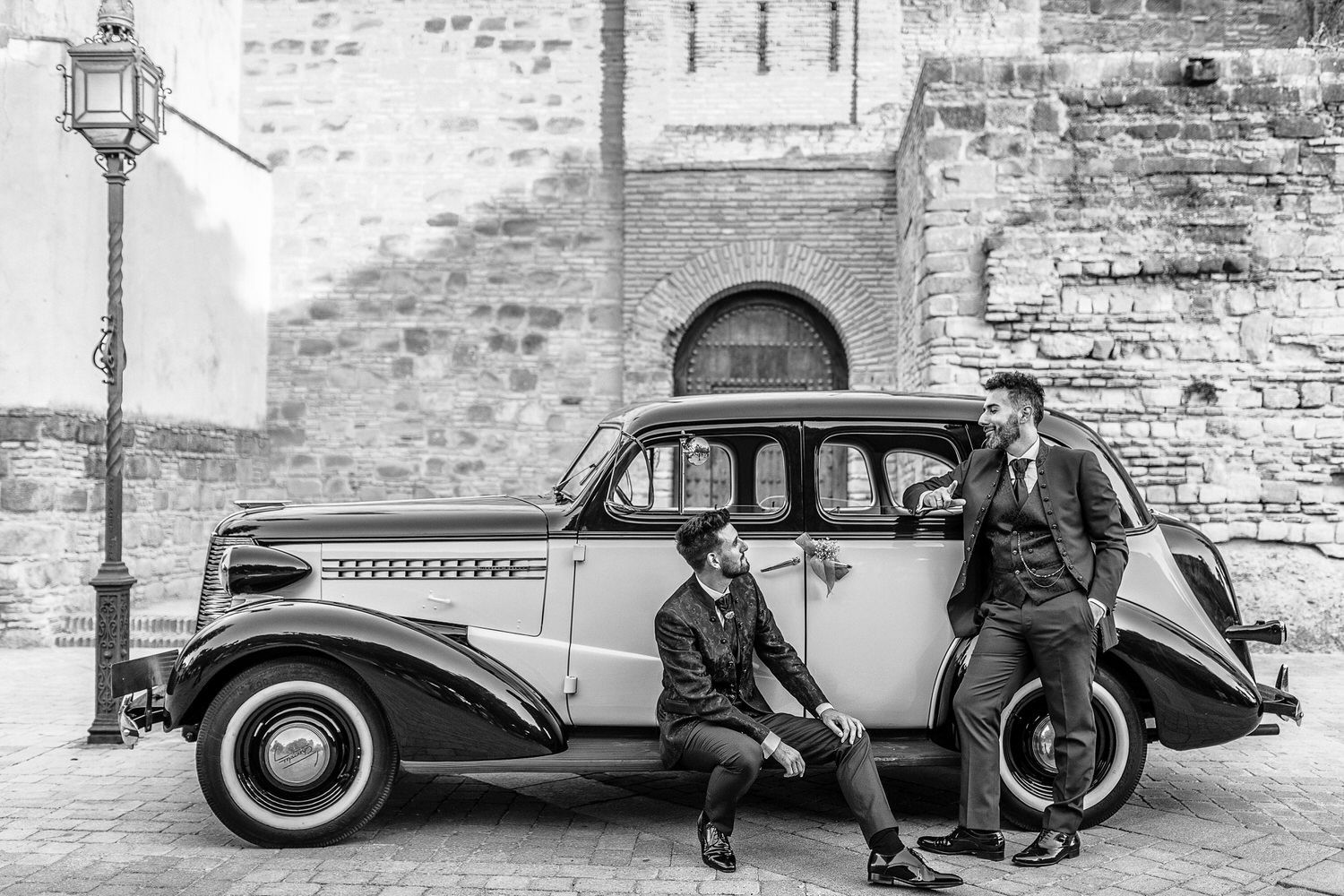 Dos hombres de traje posan con un coche clásico frente a un edificio de piedra.
