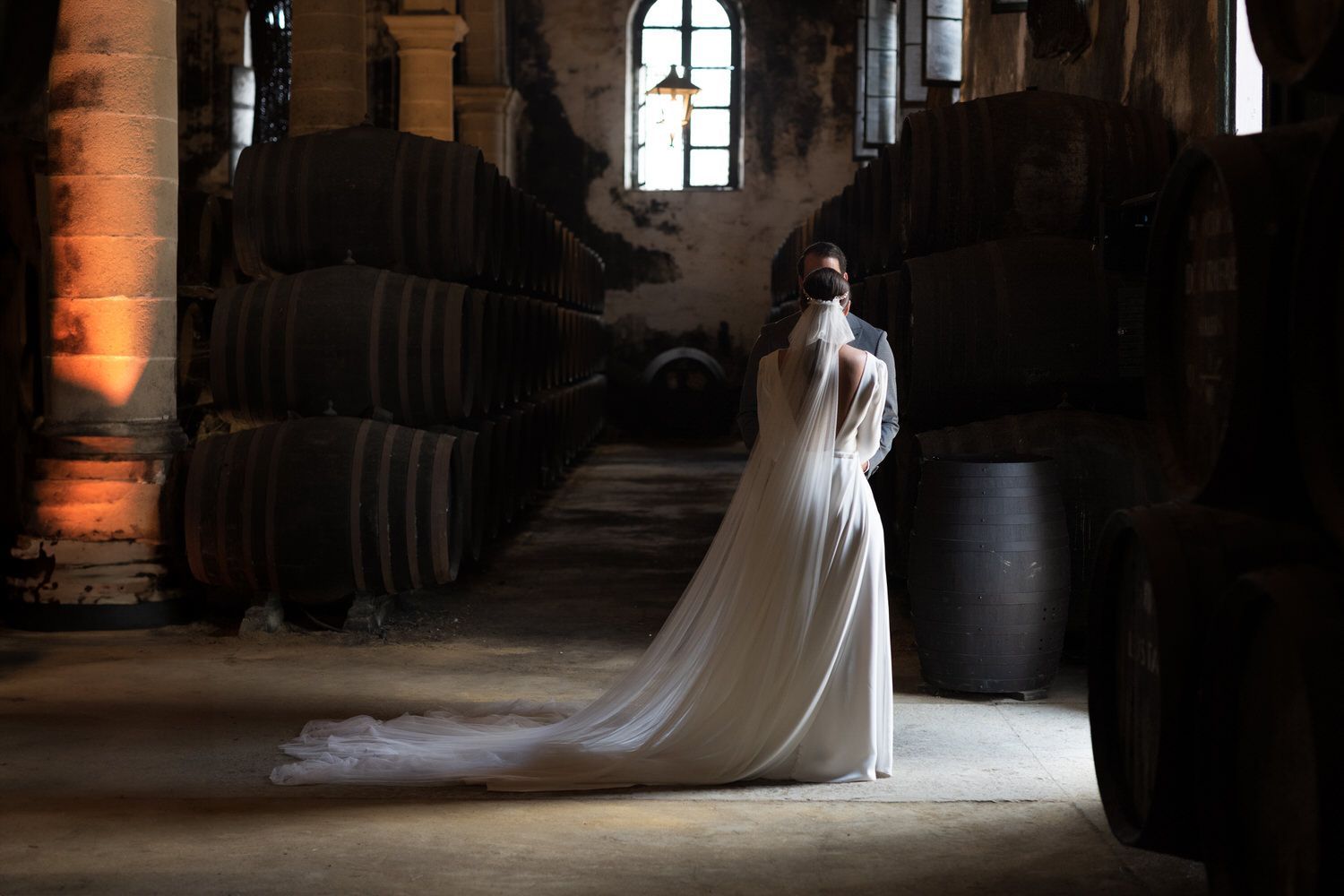 Novia con un vestido de novia con una larga cola, de espaldas en una habitación poco iluminada con barriles.