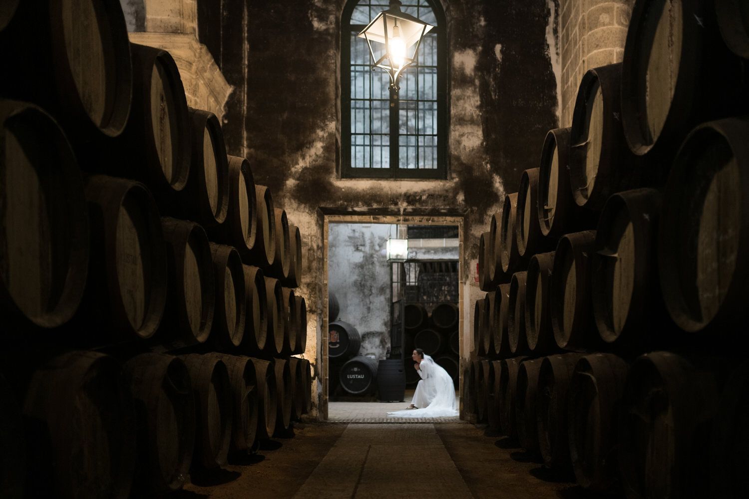 Una mujer con vestido blanco se arrodilla en una bodega, con barriles alineando las paredes. 