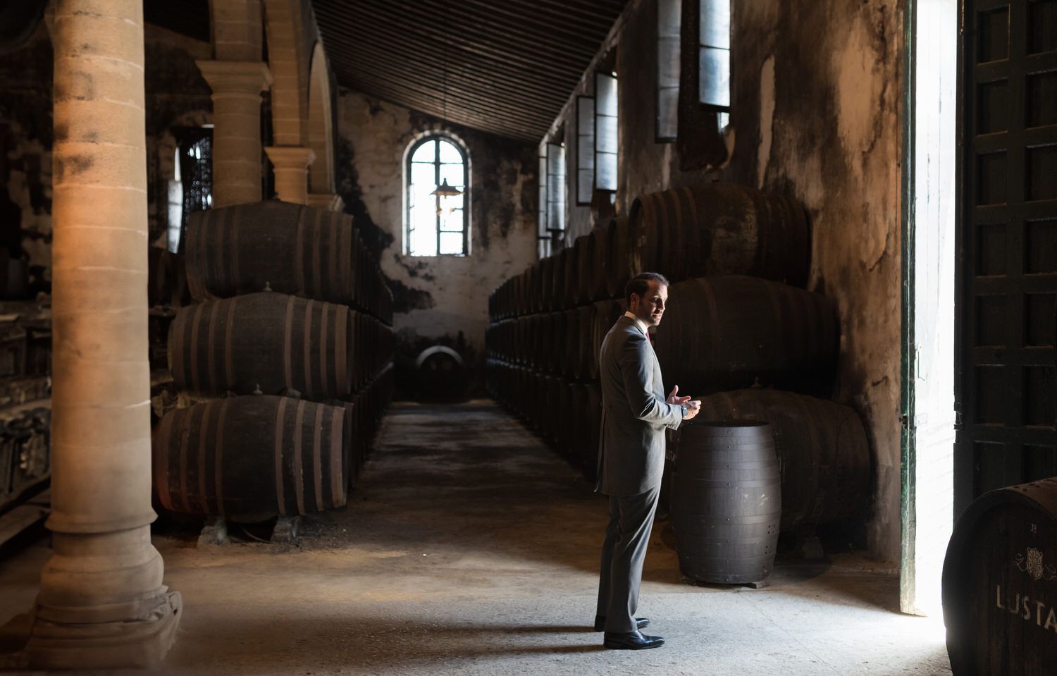 Hombre de traje de pie en una bodega poco iluminada con grandes barriles apilados contra las paredes.