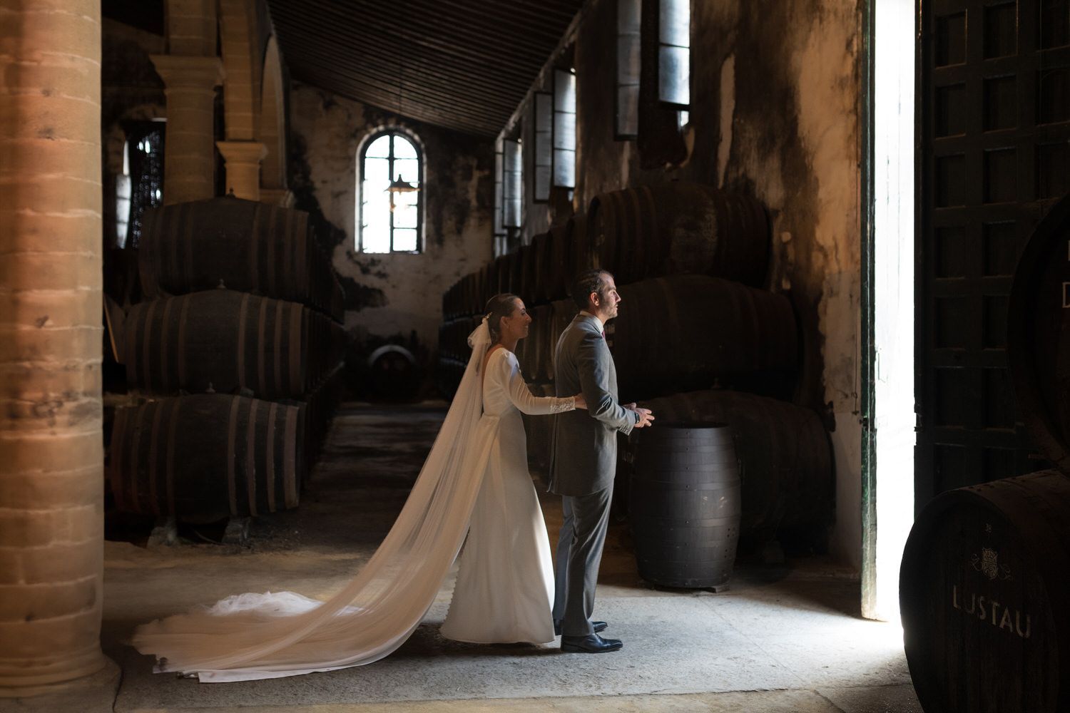 Los novios están de pie en una bodega, mirando hacia una puerta abierta. Un largo velo se extiende tras la novia.