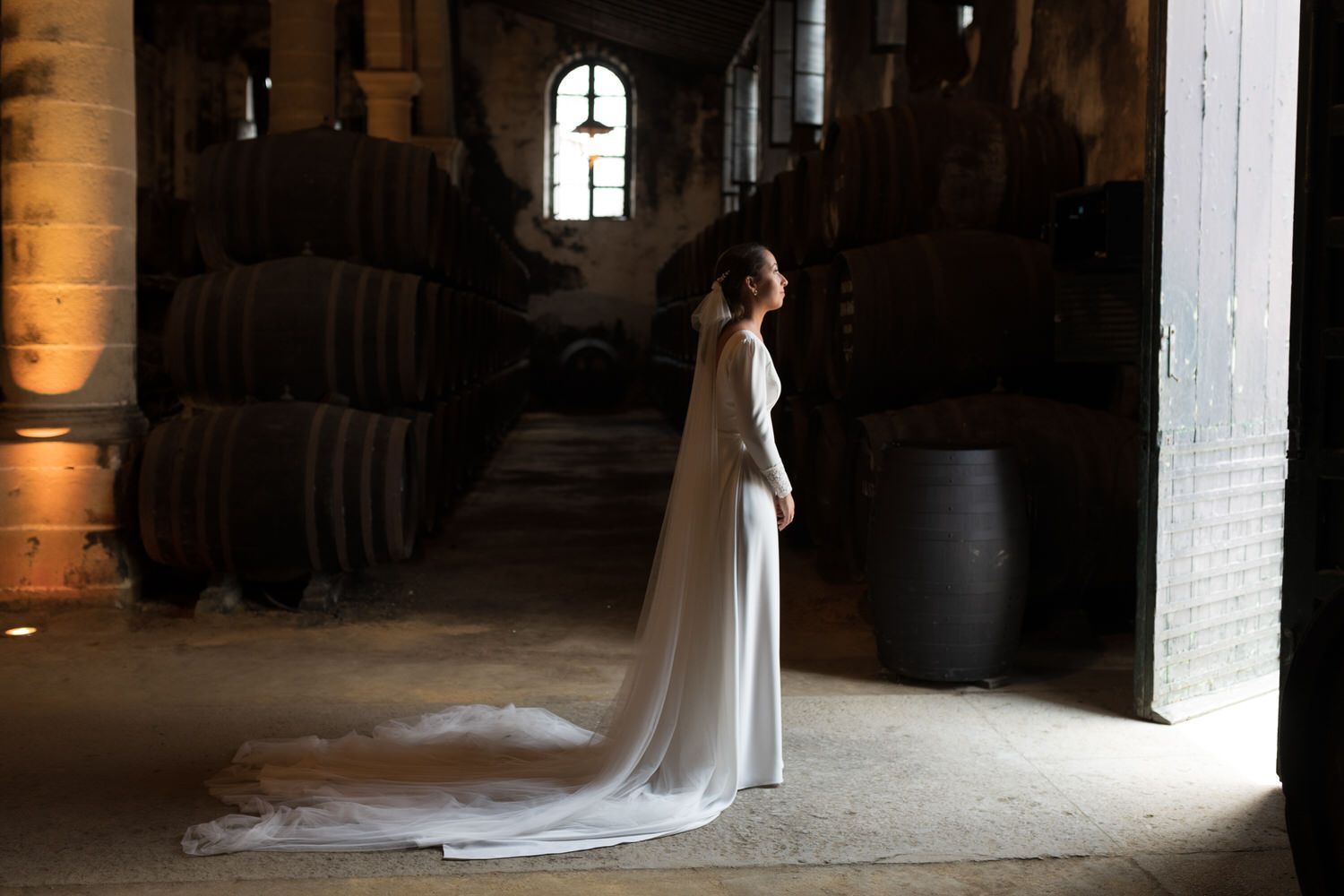 Una mujer con un vestido de novia blanco se encuentra en un sótano poco iluminado con barriles.