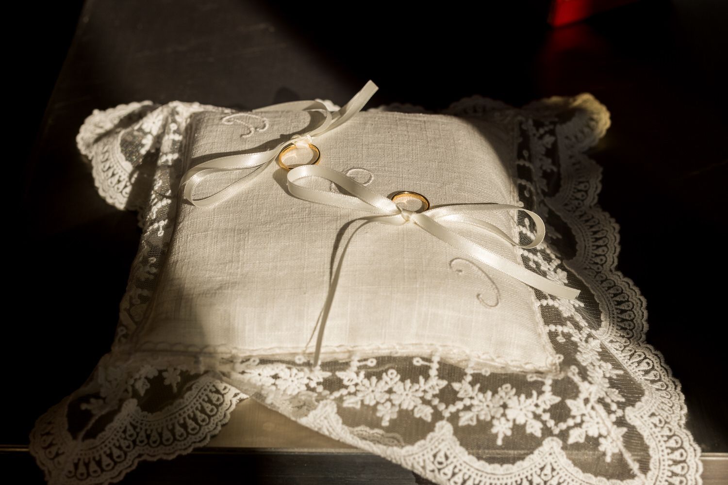 Anillos de boda sobre una almohada blanca con bordes de encaje, sostenidos con cintas blancas, bajo la brillante luz del sol.