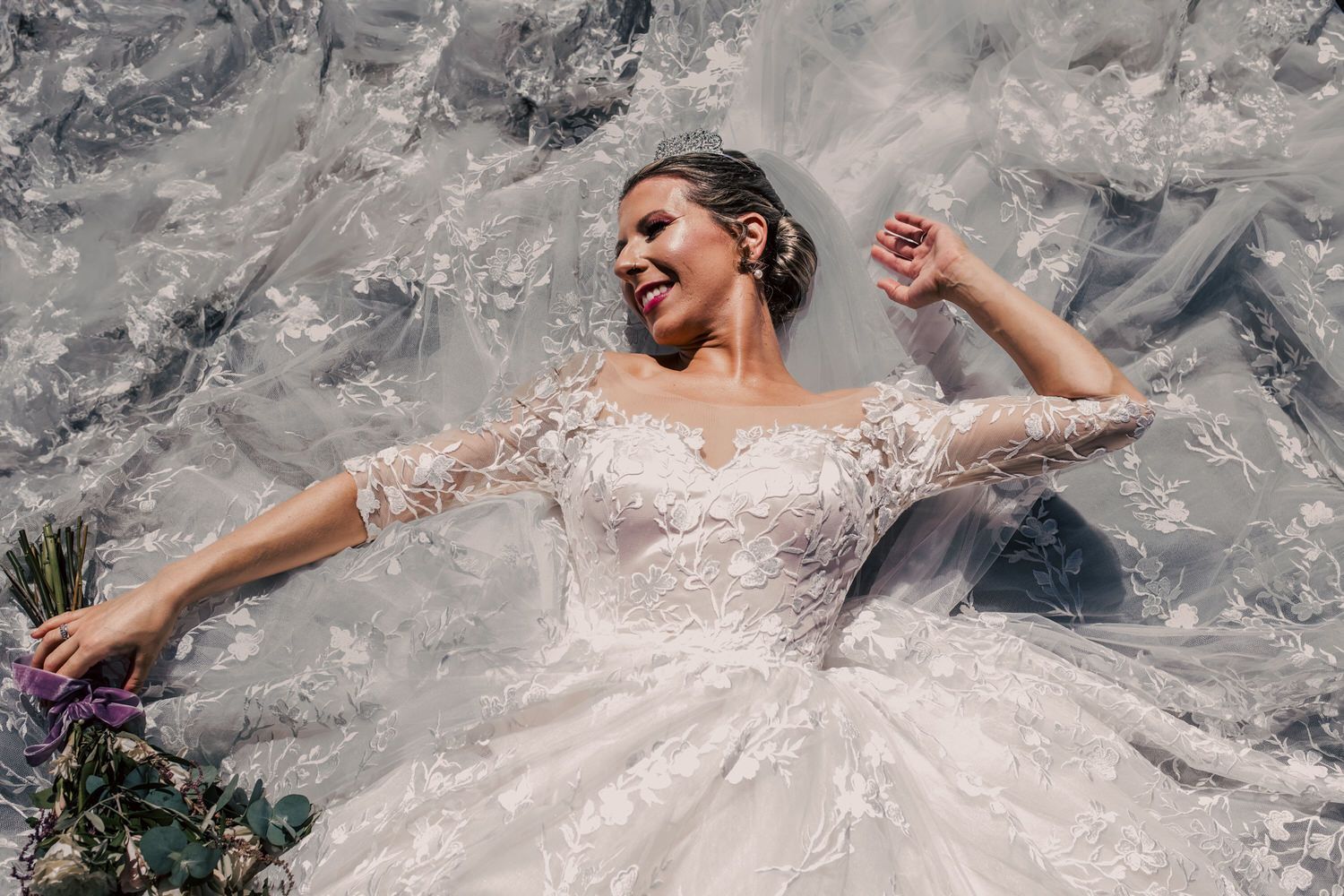 Novia con vestido de novia de encaje blanco, sonriendo, recostada sobre el vestido. Sosteniendo flores al aire libre.