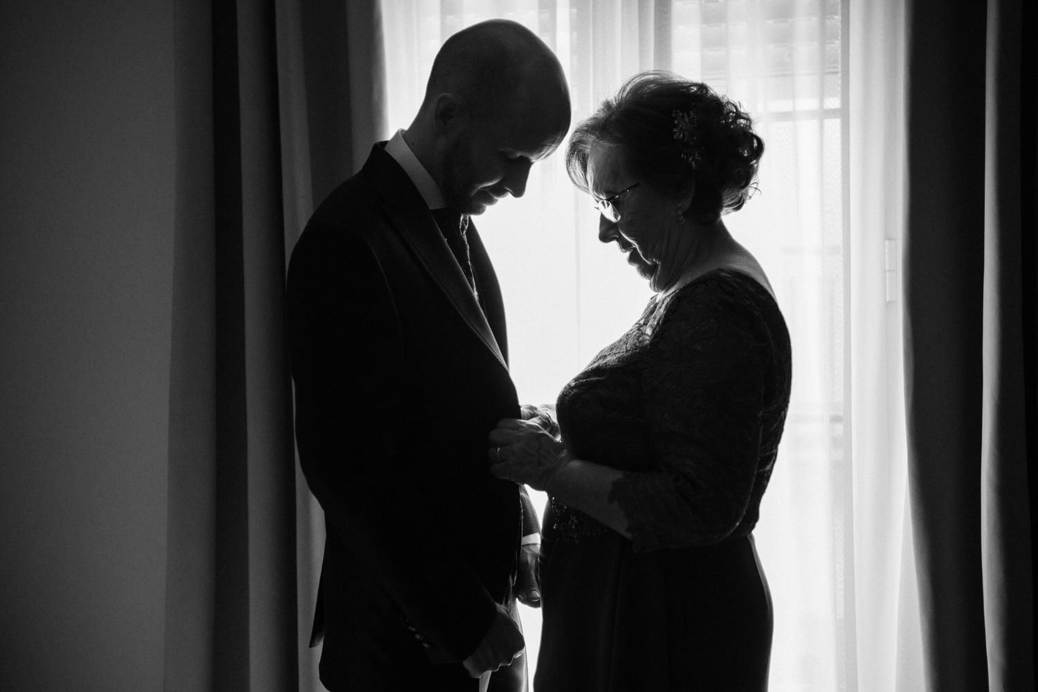Silueta de una persona con traje y una persona con vestido cerca de una ventana, con la cabeza inclinada.