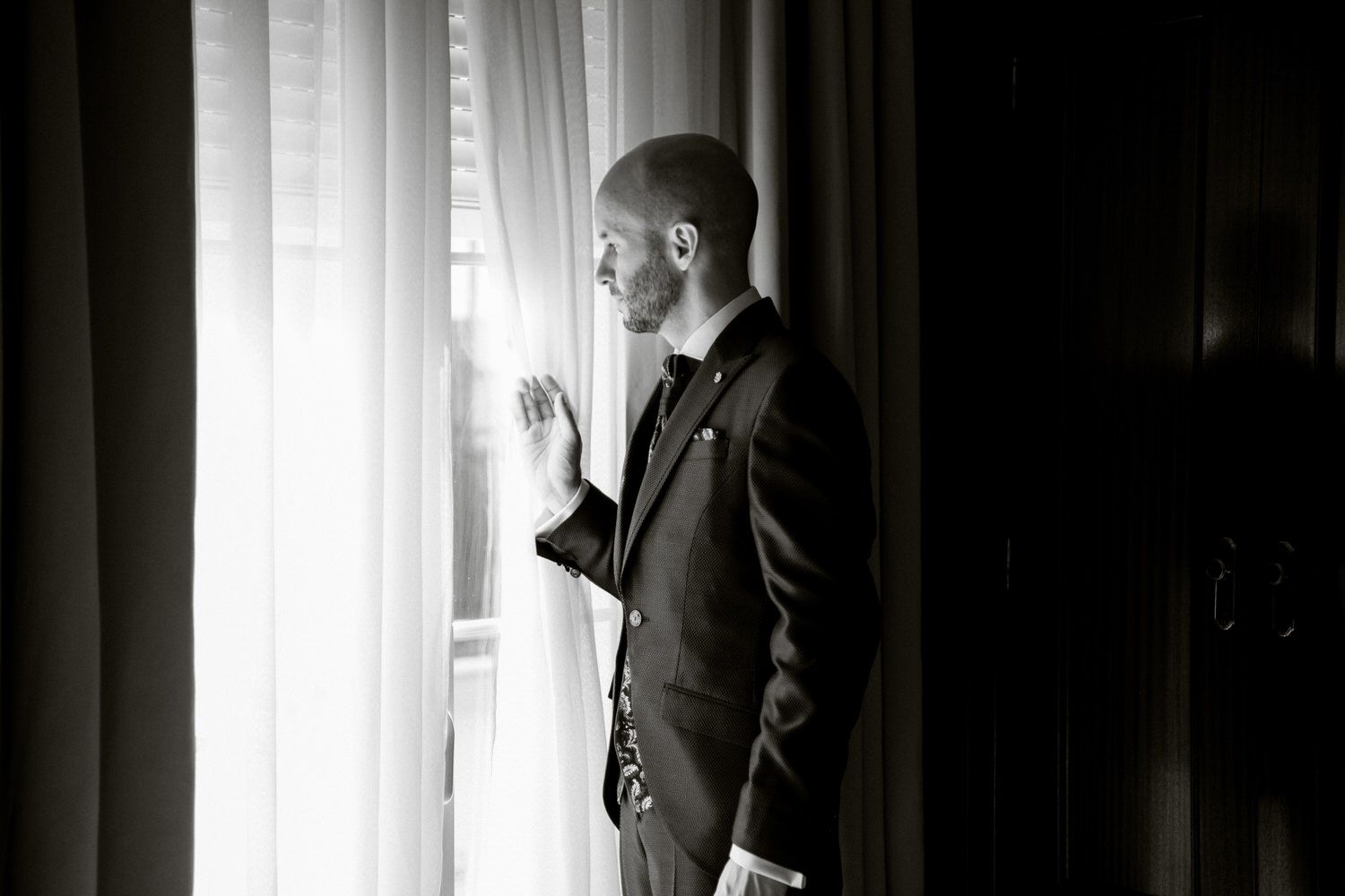 Hombre de traje mirando por una ventana, con la mano en alto. Foto en blanco y negro, interior bien iluminado.