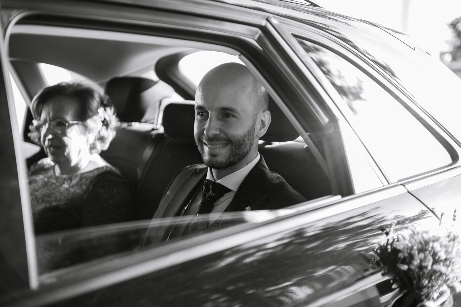 Un hombre con traje sonríe desde el asiento trasero de un automóvil, una mujer a su izquierda y flores visibles.