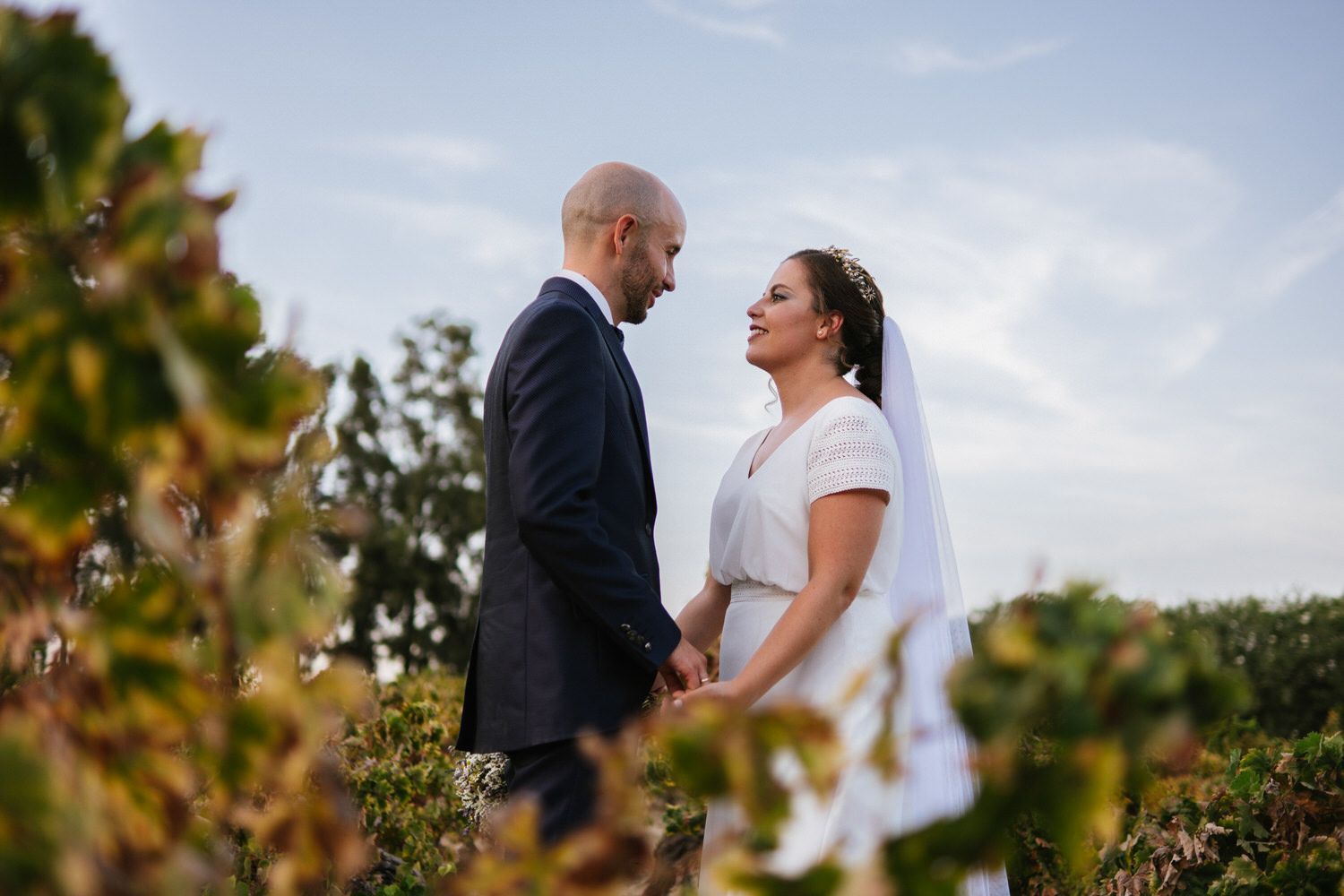Una pareja vestida de novia se toma de las manos y se mira en un viñedo, soleado y con un cielo azul.
