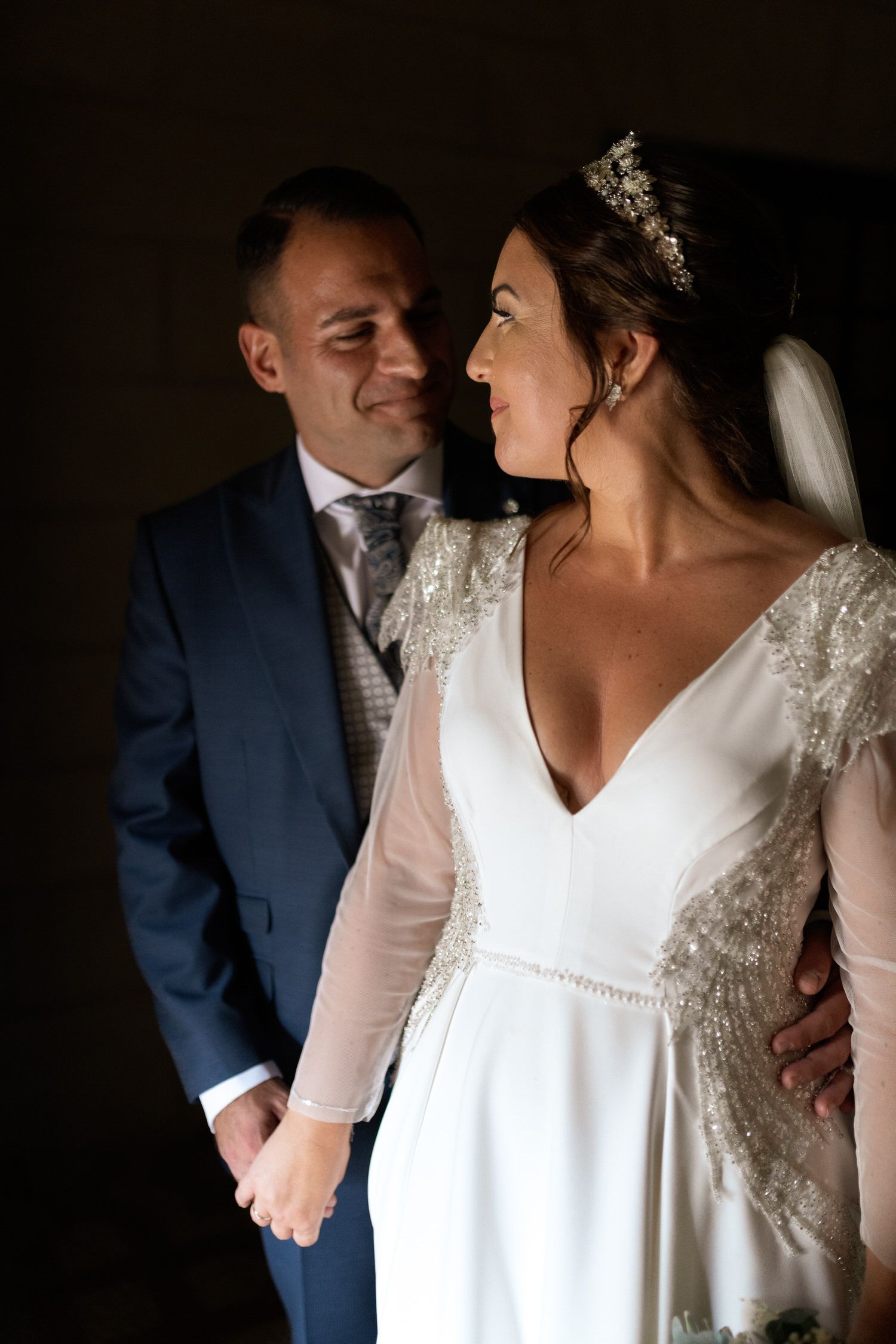 Los novios se miran de la mano. La novia lleva vestido blanco y velo, y el novio, traje azul.