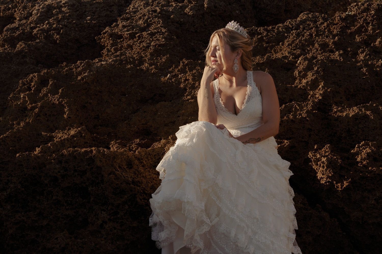 Novia con un vestido blanco y falda con volantes, sentada y mirando hacia un lado con la mano cerca de la cara.