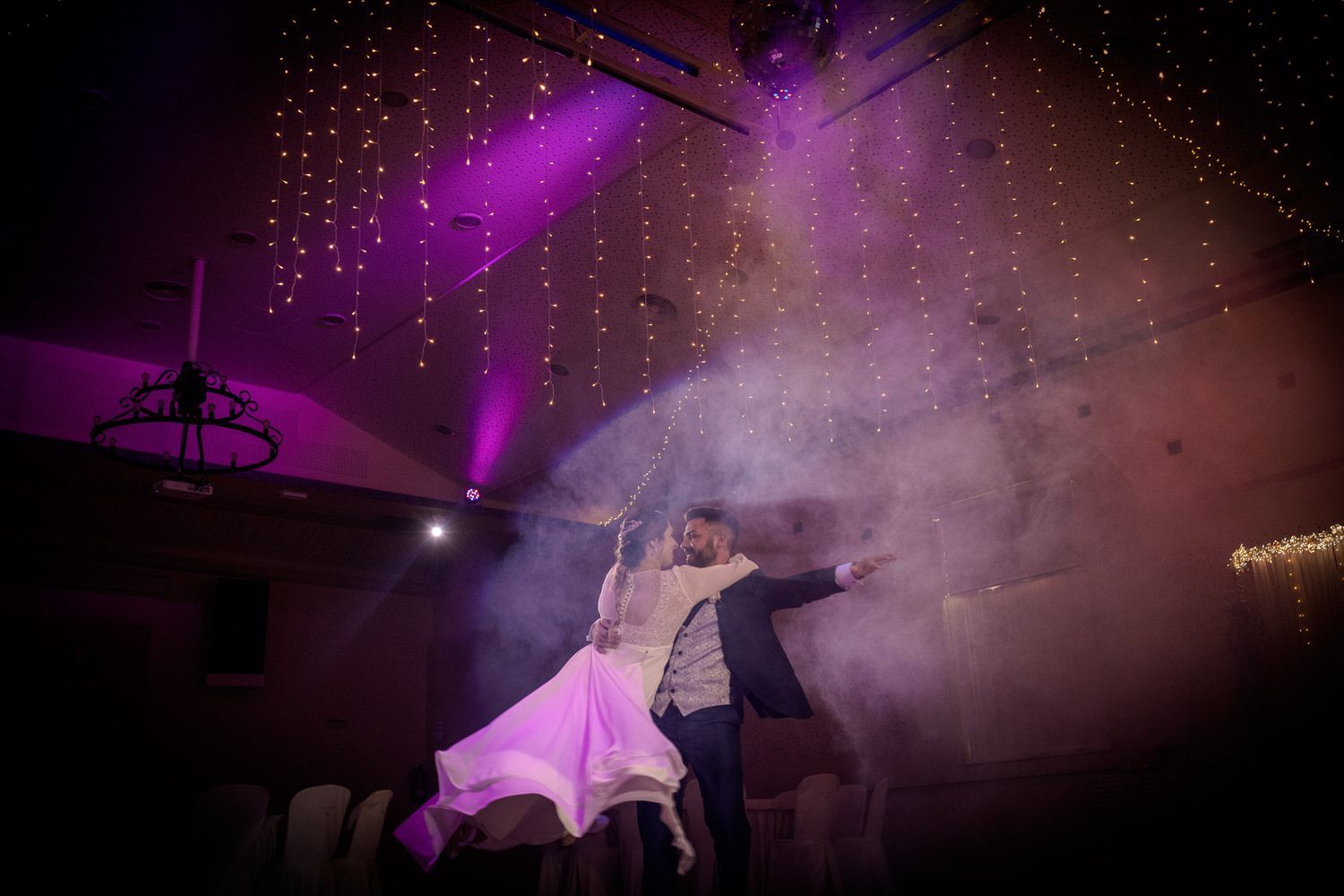 Pareja bailando bajo una luz violeta, rodeada de humo y con luces de colores colgantes en un local.