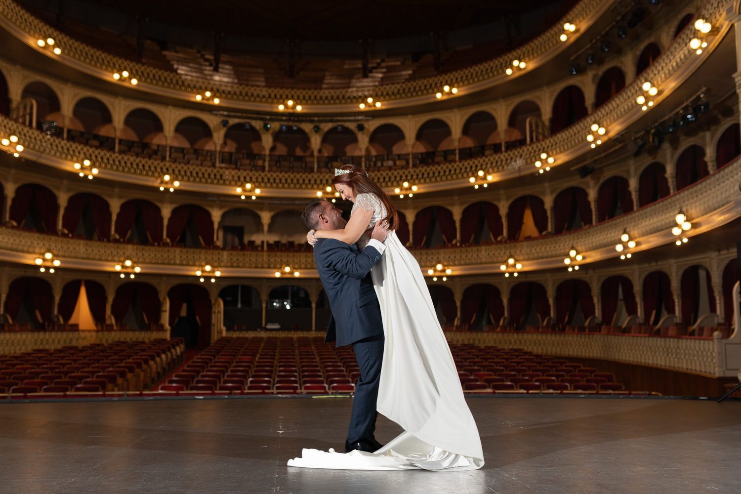 Los novios se abrazan en un escenario con gradas. El novio levanta a la novia en el aire.