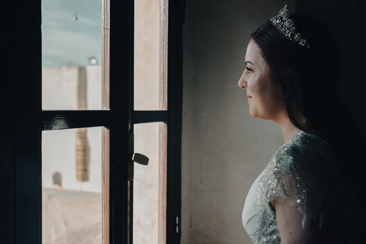 Una novia con tiara mira por la ventana, de perfil. El entorno es probablemente al aire libre, en una habitación oscura.