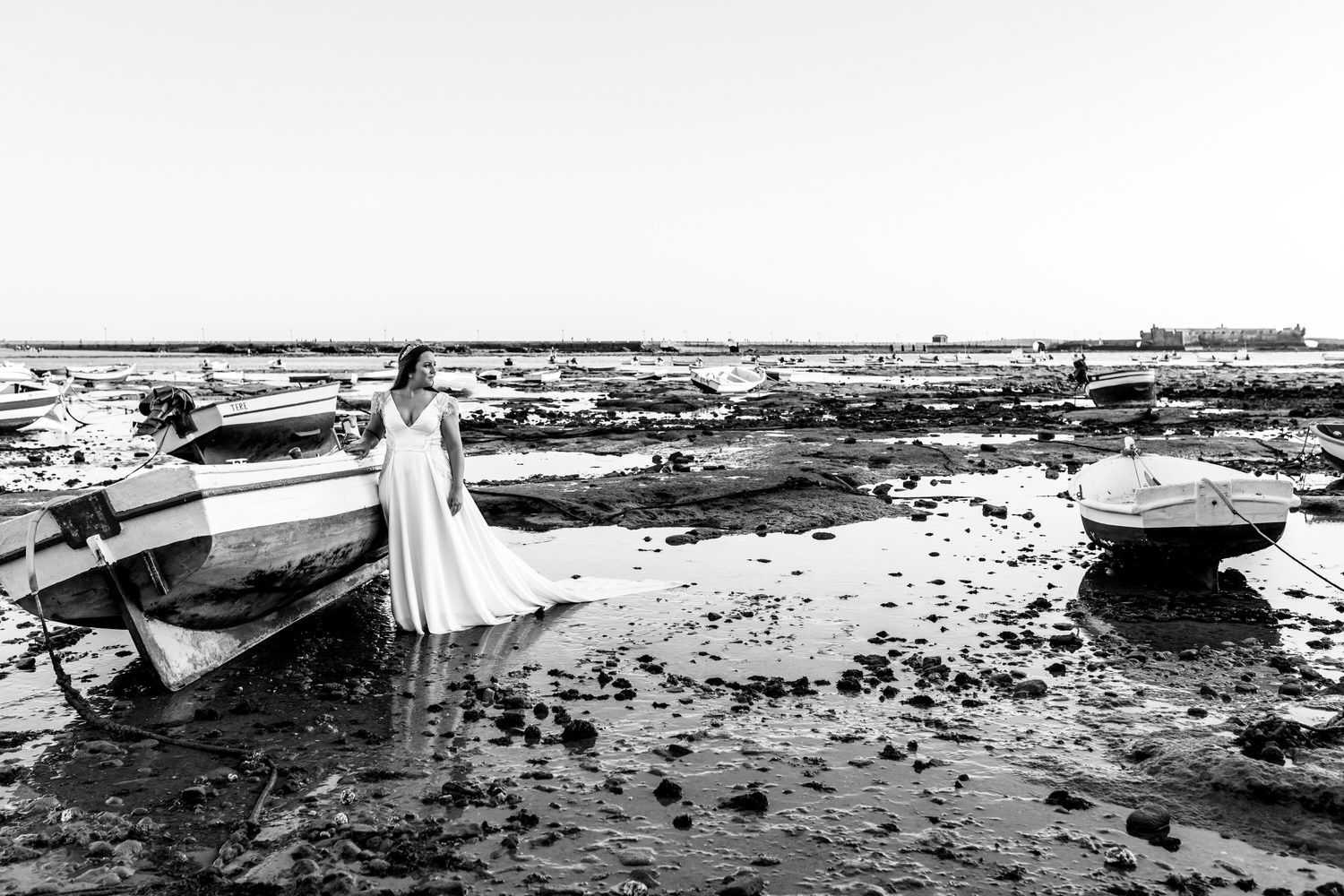 Mujer con vestido de novia se encuentra en una orilla fangosa cerca de barcos varados; blanco y negro.