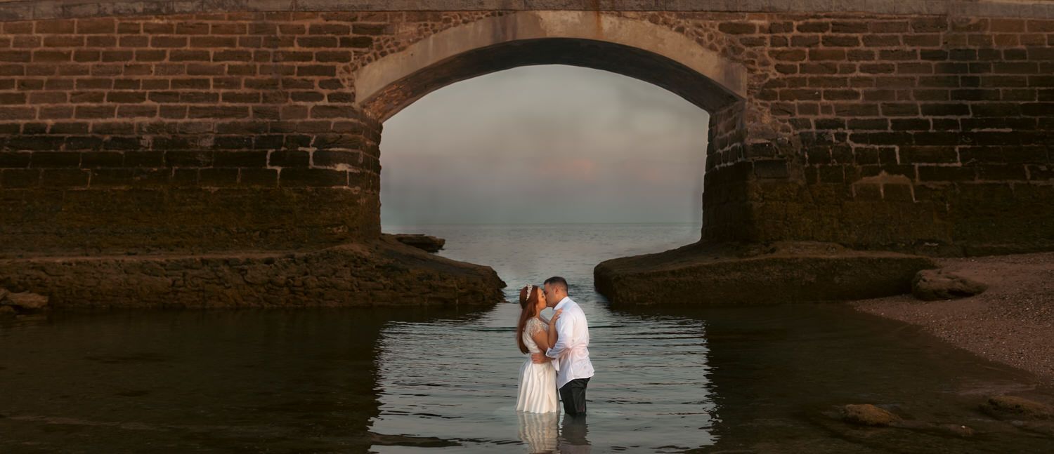 Pareja besándose bajo el arco de un puente de ladrillo, de pie en aguas poco profundas.