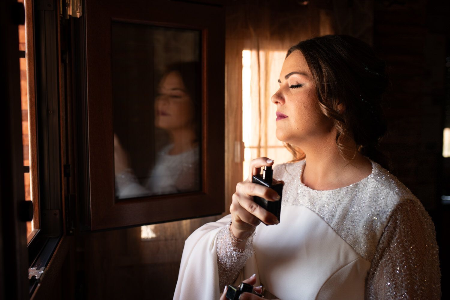 La novia se perfuma frente a una ventana, con su reflejo visible. Lleva un vestido blanco.