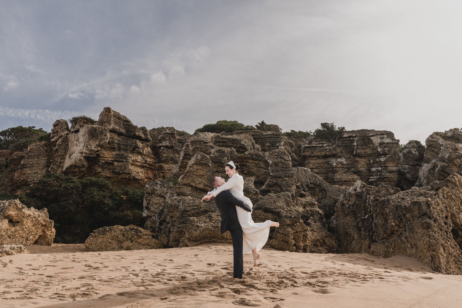 El novio levanta a la novia en una playa con formaciones rocosas bajo un cielo nublado.