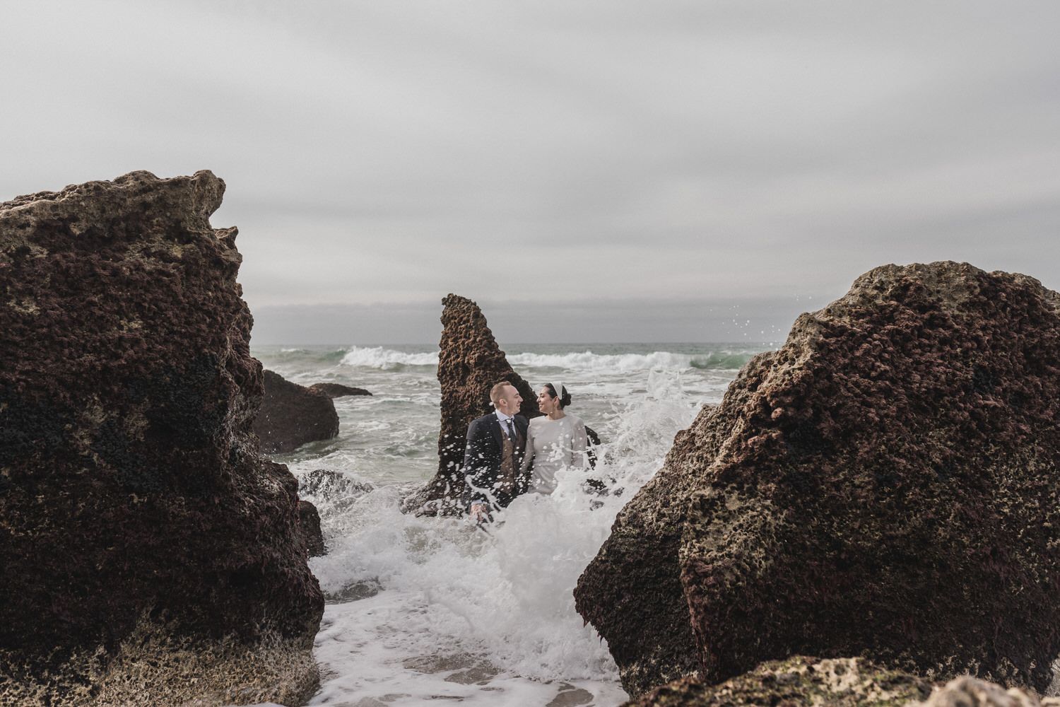 Una pareja vestida de manera formal se encuentra frente a las olas del océano, entre las rocas; cielo nublado.