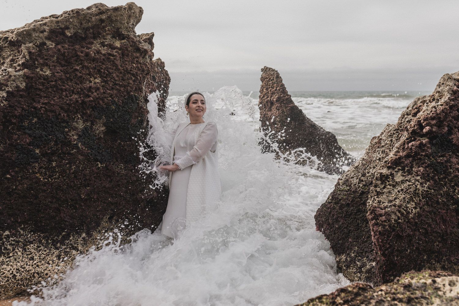 Una mujer vestida de blanco se encuentra frente a las olas del océano que se estrellan entre formaciones rocosas.