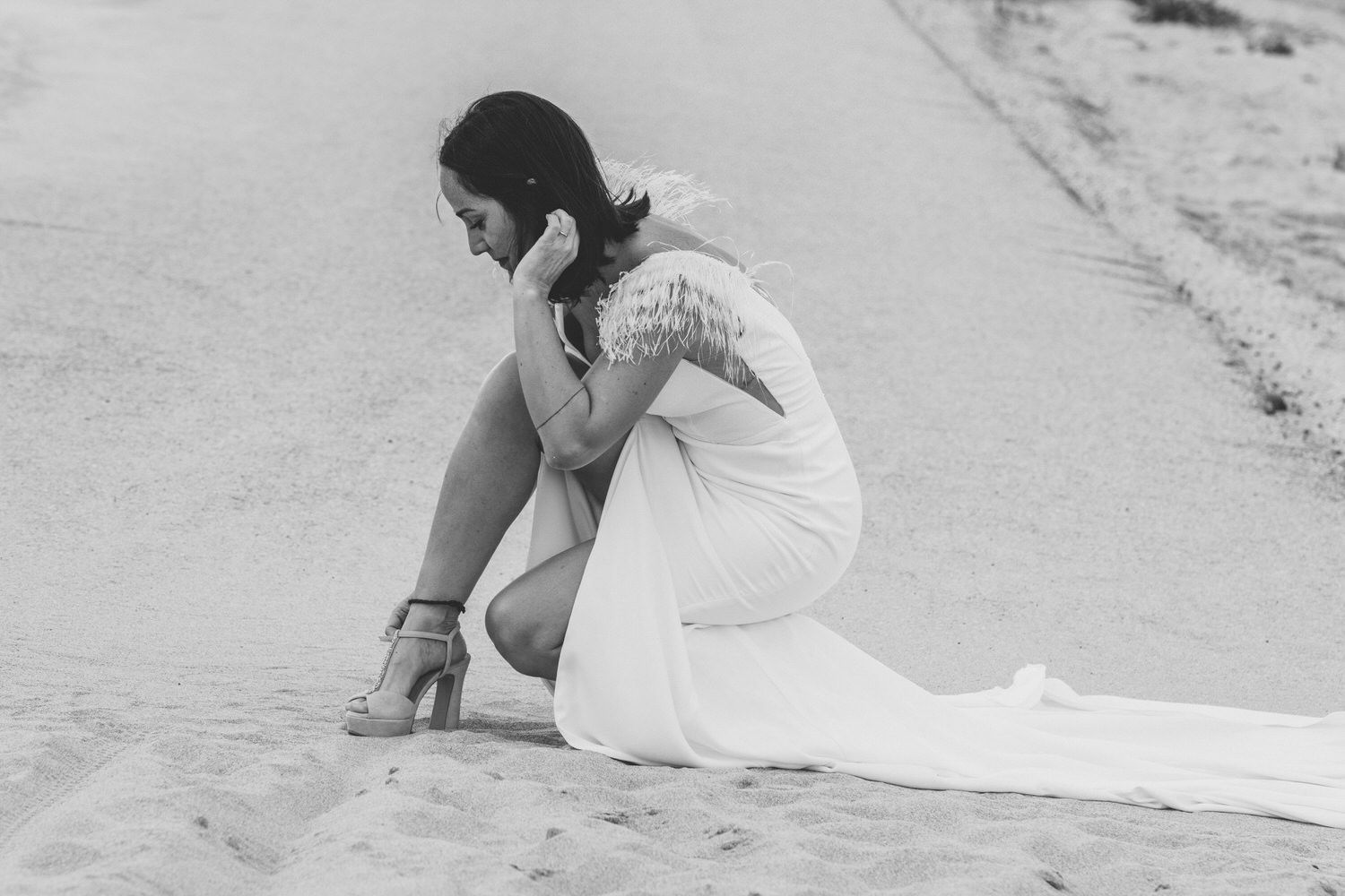 Mujer con vestido de novia, arrodillada en un camino de arena, apoyando la cabeza en la mano.