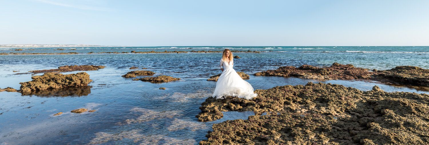 Una novia con un vestido blanco está parada sobre rocas en aguas poco profundas junto al océano, bajo un cielo azul.