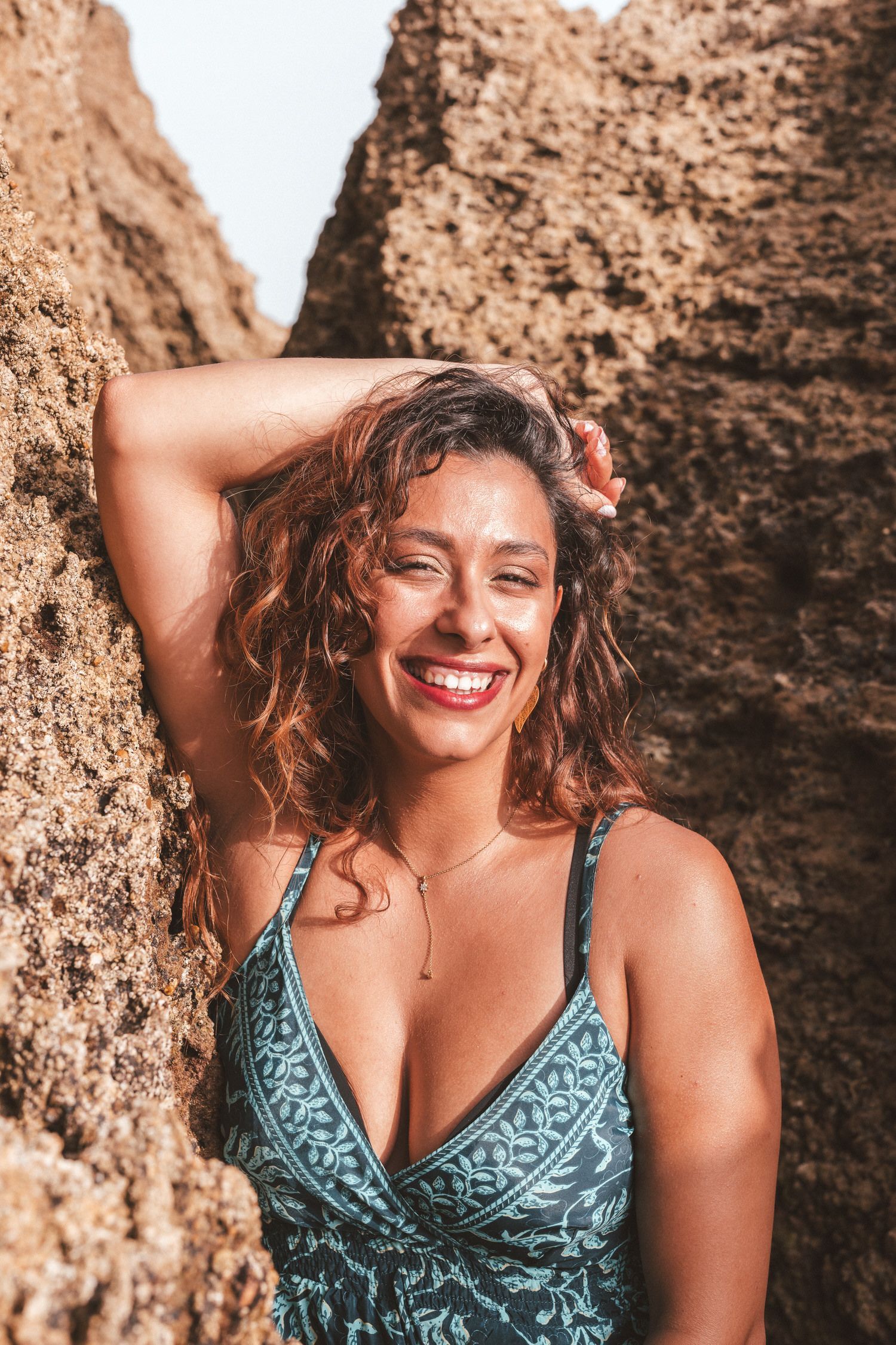 Mujer con vestido azul, apoyada contra una pared rocosa, sonriendo brillantemente.