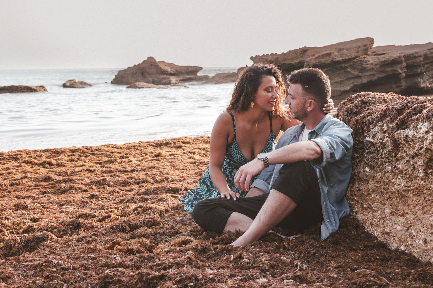 Una pareja sentada cerca en una playa cubierta de algas, mirándose. El océano y las rocas al fondo.