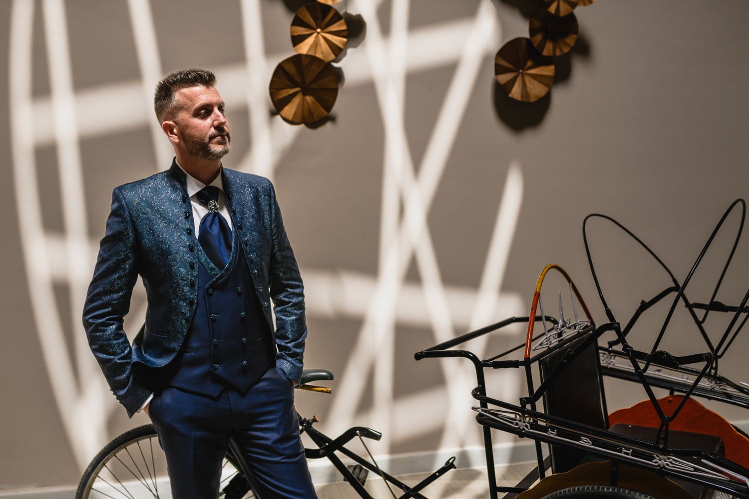 Un hombre con traje azul posa junto a una escultura de bicicleta deconstruida. Fondo neutro con arte mural y sombras.