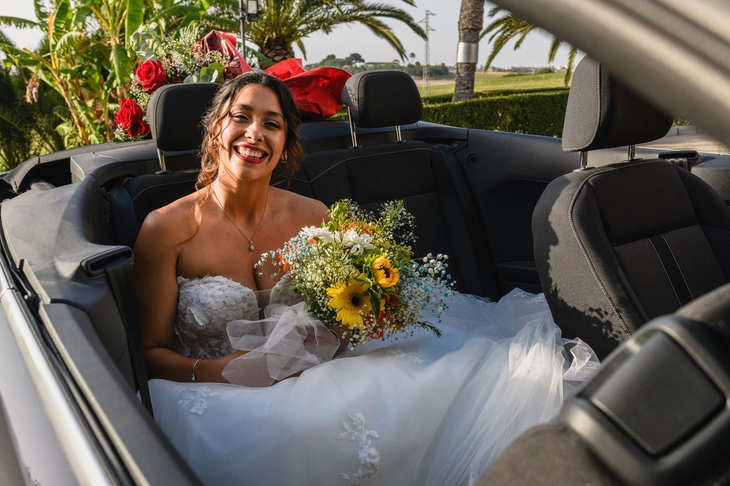 Novia con vestido de novia blanco con ramo, sonriendo en coche descapotable.