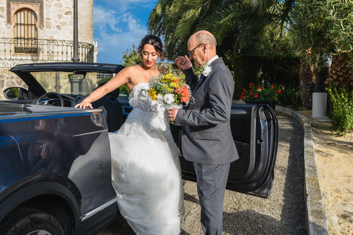 Novia saliendo del coche, sosteniendo un ramo, asistida por un hombre de traje, exterior cerca del edificio.
