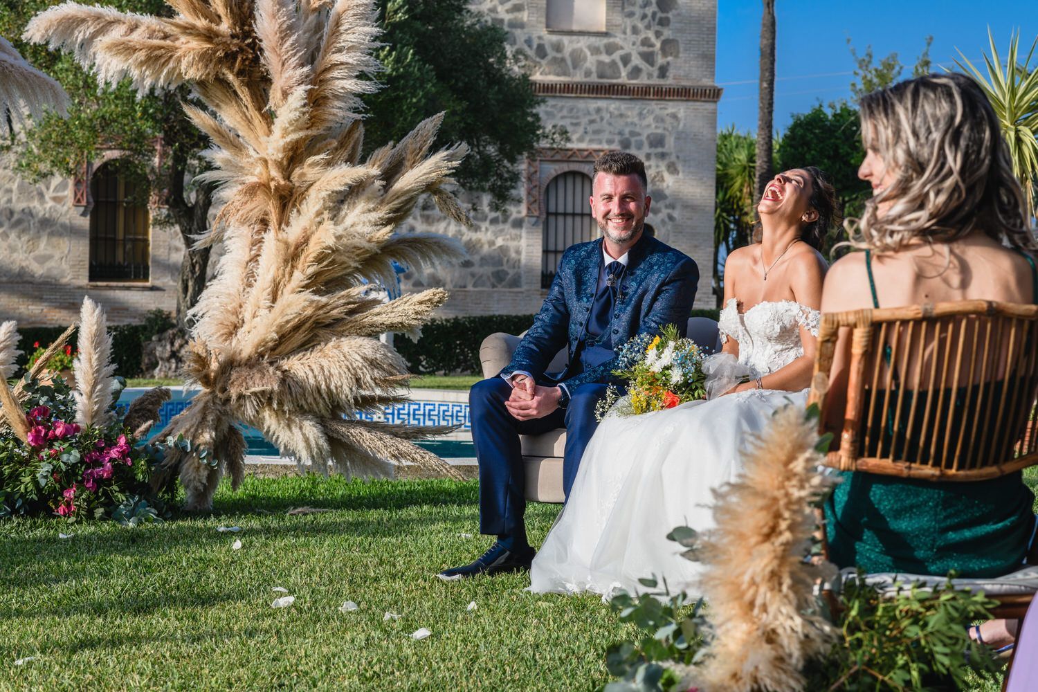 El novio y la novia sentados al aire libre durante una ceremonia nupcial. La novia ríe, sosteniendo el ramo. Otros sentados.