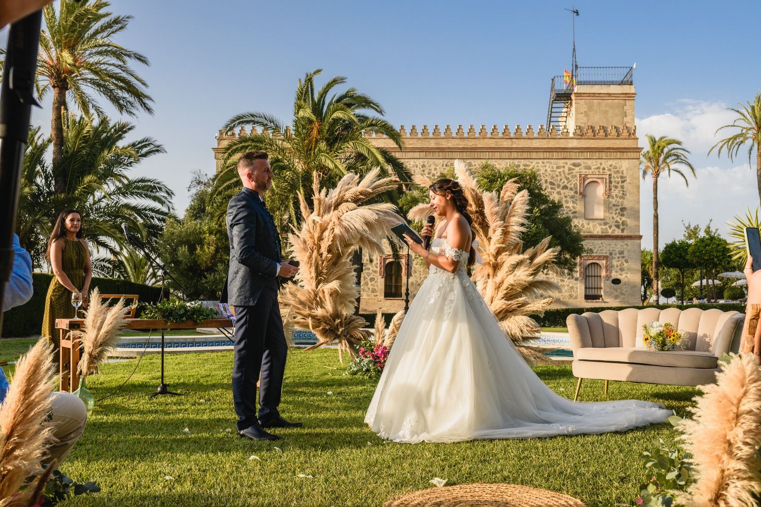 Ceremonia de boda al aire libre; la novia lee sus votos al novio, día soleado, edificio histórico al fondo.