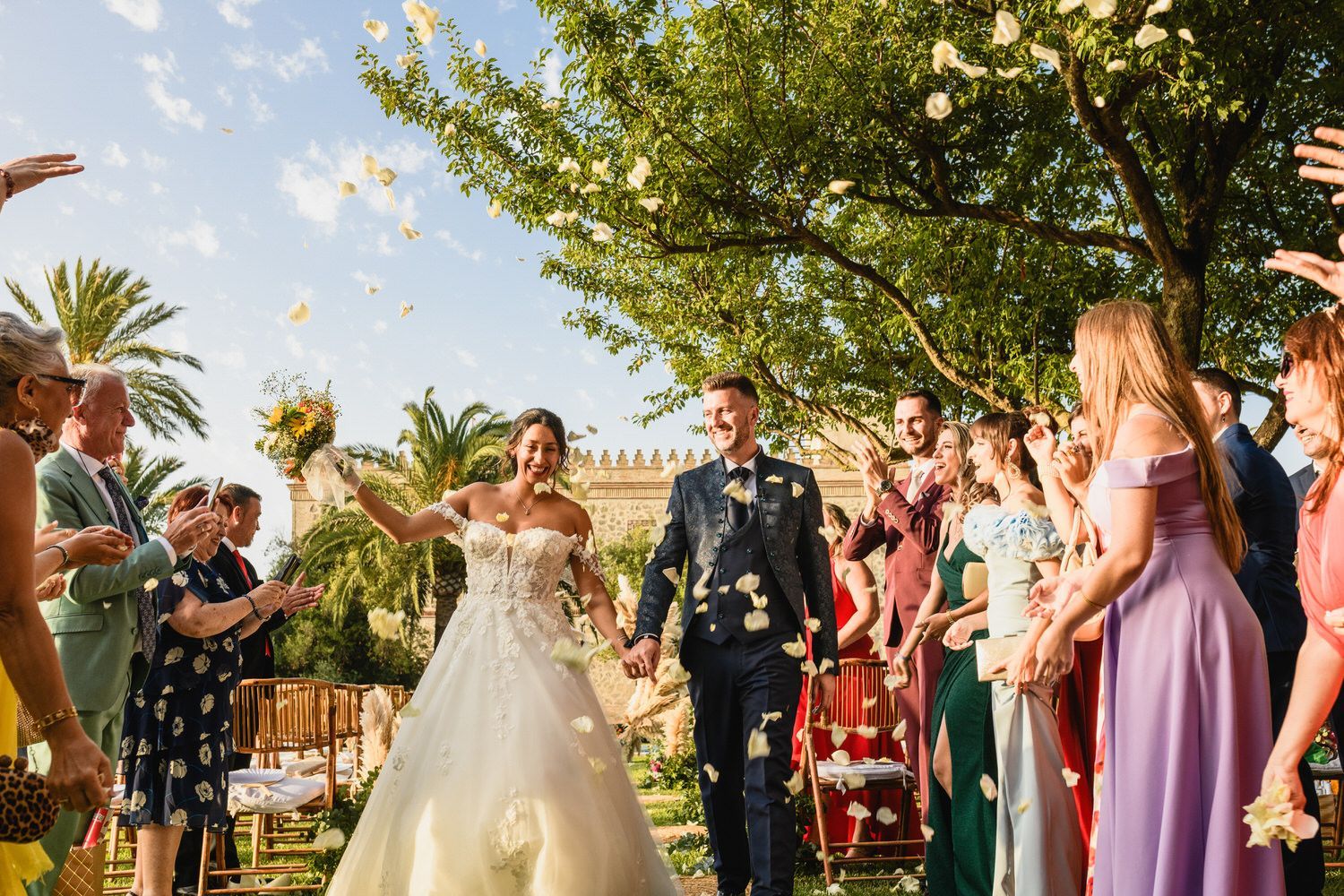 Los recién casados ​​caminan entre una lluvia de pétalos de flores, mientras los invitados vitorean; boda al aire libre.