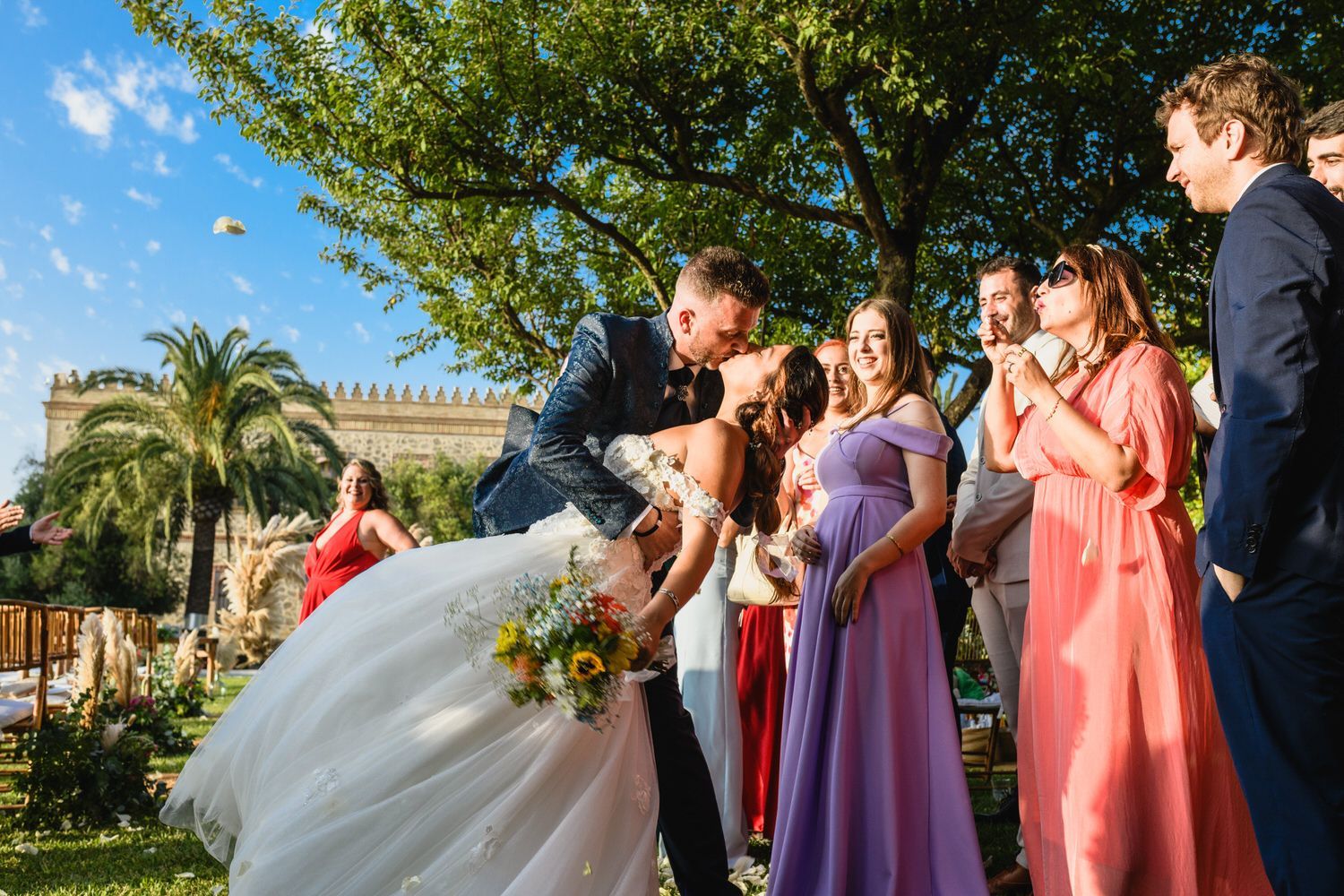 Los novios se besan en una ceremonia de boda, rodeados de invitados en un entorno al aire libre con cielo azul.