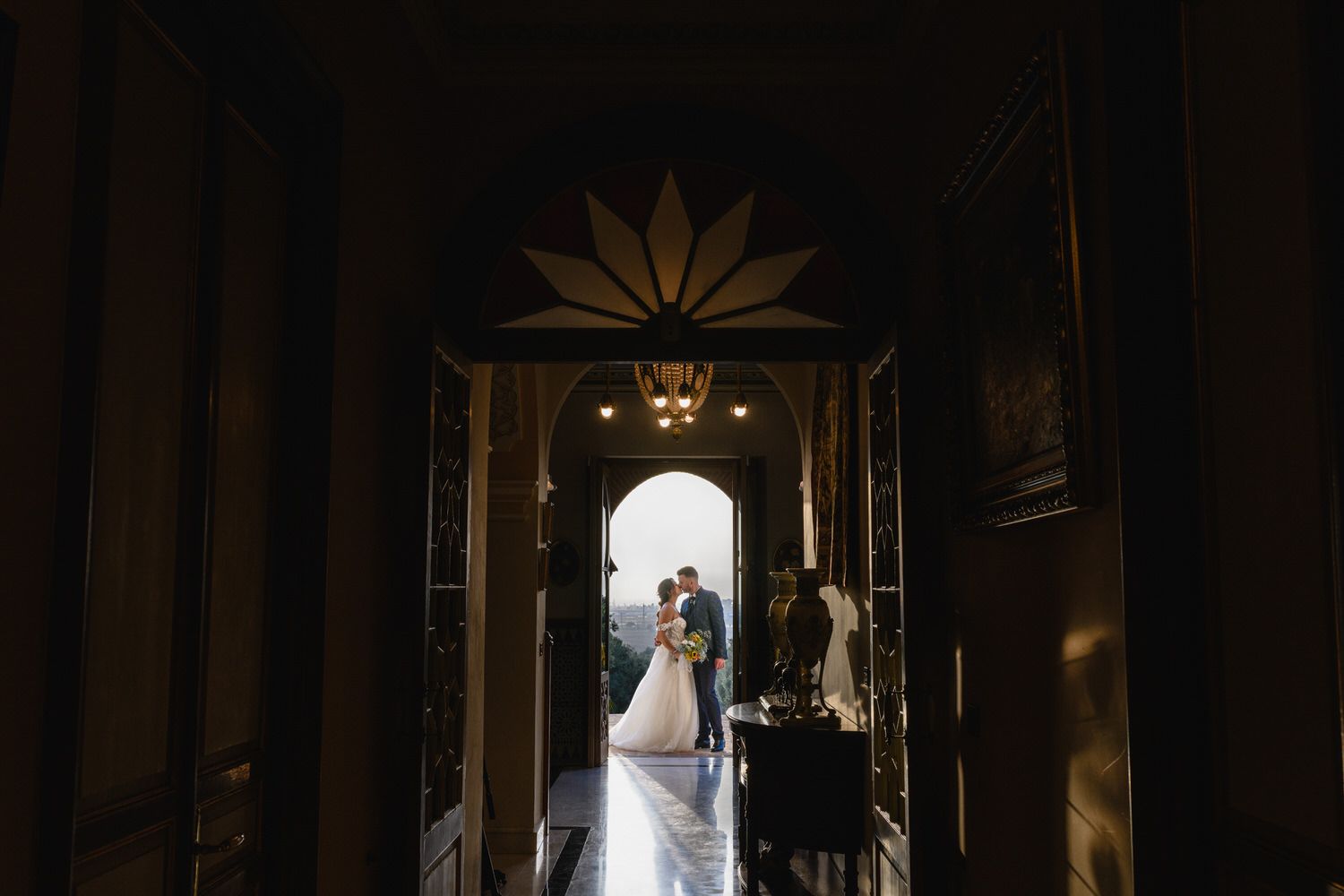 Una pareja vestida de novia se abraza en una puerta iluminada por el sol, con un pasillo oscuro enmarcando la escena.