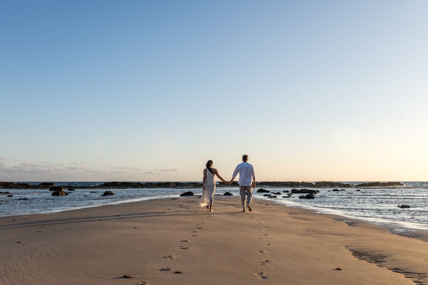Pareja tomada de la mano, caminando por un sendero de arena hacia el horizonte, agua y rocas a ambos lados.