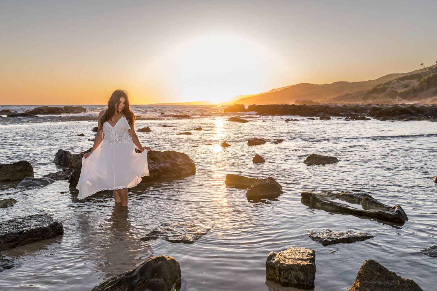 Mujer con vestido blanco caminando en el océano, sosteniendo el vestido, puesta de sol sobre el agua.