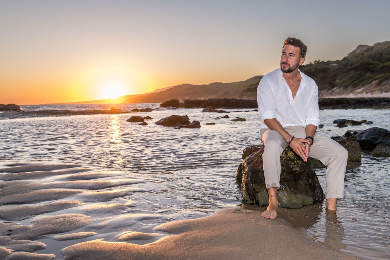 Hombre sentado en una roca en una playa, mirando la puesta de sol; camisa blanca, pantalones beige.